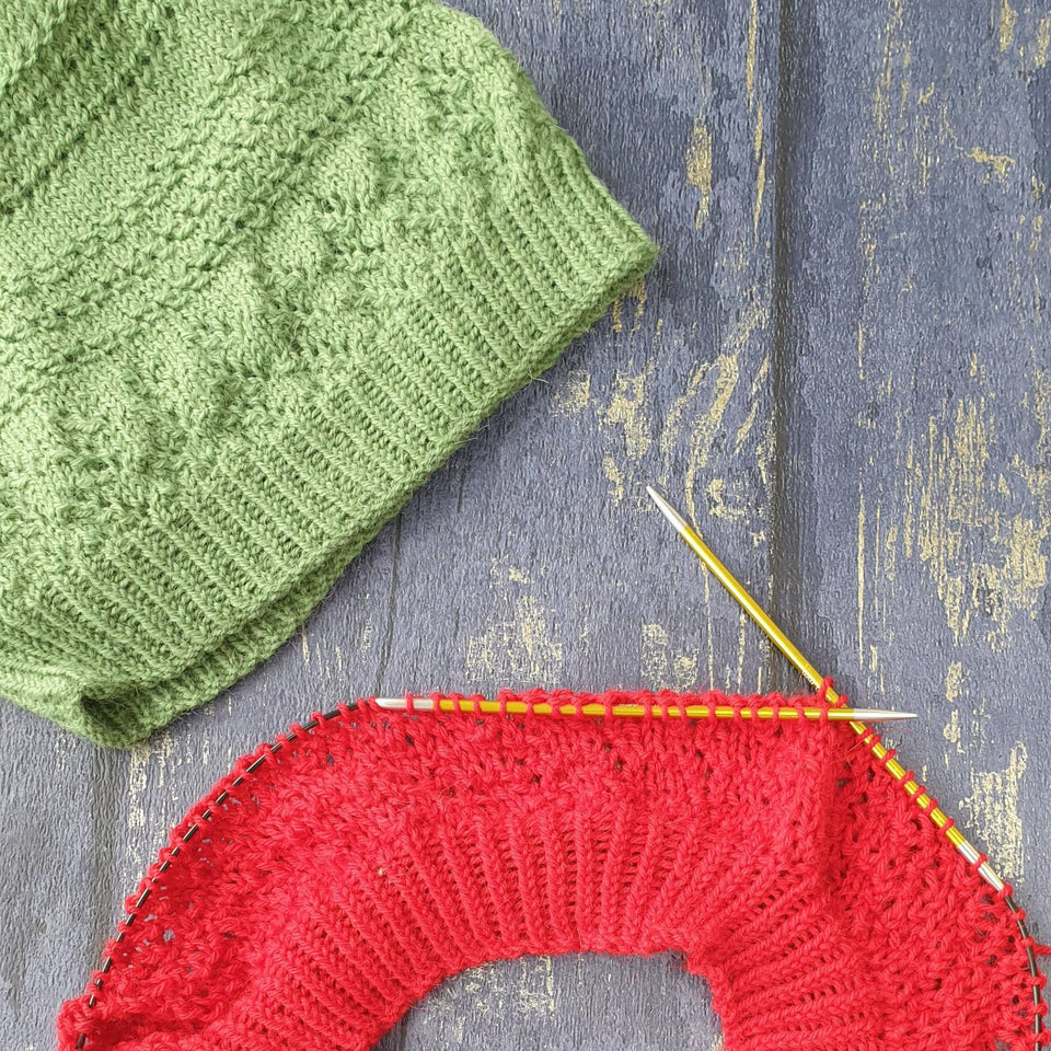 green textured hat in the left corner and red textured hat on circular knitting needles, on a blue grey wood background